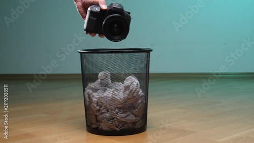 Human hand dropping an outdated professional digital single lens reflex camera into a metal trash bin filled with crumpled waste paper, symbolizing obsolescence and electronic waste disposal
