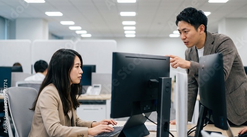 Two people are working together in a modern office. A woman sits at her desk typing on a computer, while a man stands beside her, pointing at the screen as they discuss something.