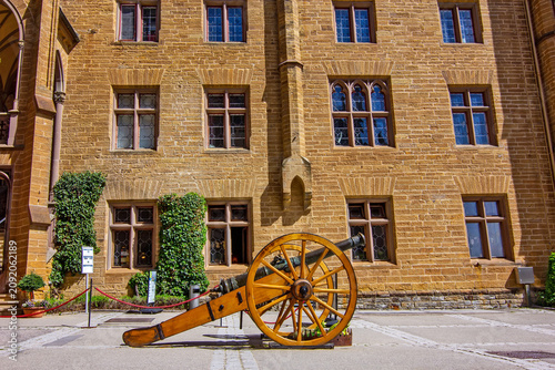 Historic courtyard at Hohenzollern Castle featuring a restored cannon, Gothic architecture, medieval buildings, and a bright blue sky in Germany.