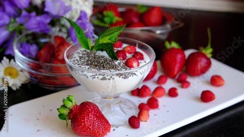 Female hand adding chia seeds to yogurt with fresh strawberries on white plate, healthy breakfast bowl with berries, nutritious superfood meal on black table with spring flowers