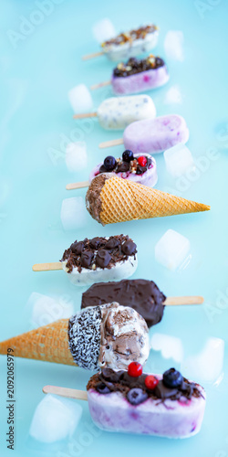 Colorful row of homemade yogurt and milk ice pops. Vanilla and chocolate decorated with nuts and frozen berries with ice cubes on bright blue background. Perfect for a summer children's birthday party
