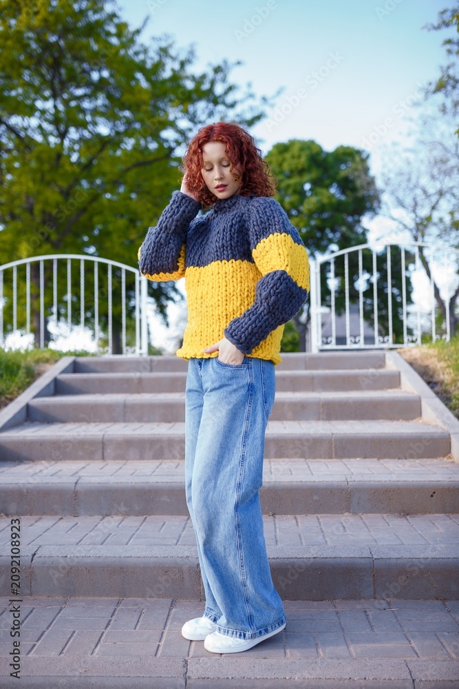 Fototapeta premium Redhead teenage girl model in chunky knit sweater and jeans on stairs background. Bulky knitwear, fashion and style concept