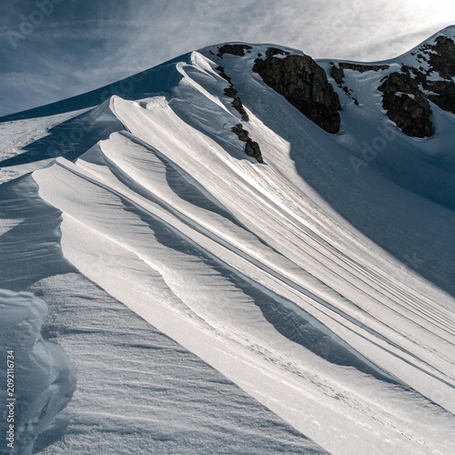 Wind sculpted snow ridge with sharp cornices and exposed dark rock, bright alpine glacier textures under pale winter sky, pristine frozen mountain wilderness in cold sunlight landscape