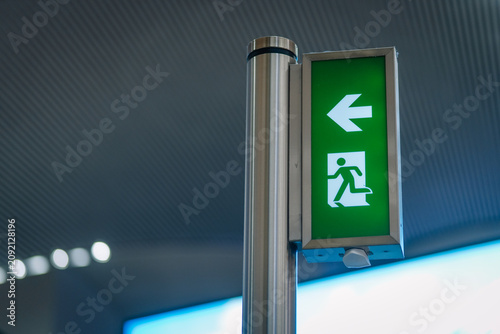 Green emergency exit sign with left arrow and running figure displayed on a metallic pole in a modern indoor setting with blurred background elements