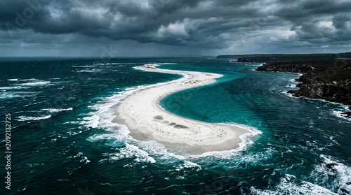 Aerial drone view of a winding white sand spit emerging from stormy ocean waters, dramatic clouds.