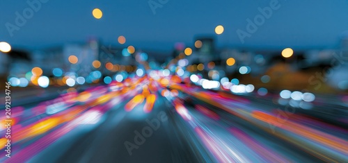 Colorful light trails flow across a bustling city street at twilight, framed by glowing orbs and a lively urban skyline.