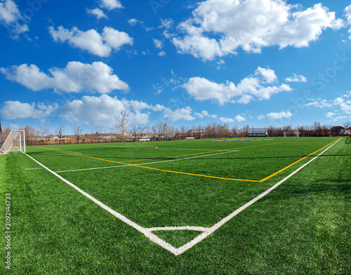 Empty soccer field with green synthetic turf, white markings, and a goal net under a bright blue sky with scattered clouds on a sunny day. Outdoor sports facility, wide angle view.