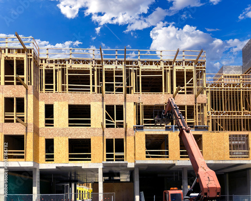 Construction site of a multi-story building featuring a wood frame structure on a concrete foundation, with a telehandler crane lifting materials against a bright blue sky.