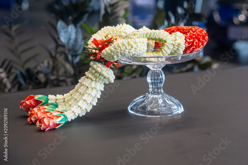 A beautiful and intricate Thai traditional flower garland (Phuang Malai) made of jasmine, crown flower, and rose petals, elegantly placed on a clear glass pedestal tray on a table.