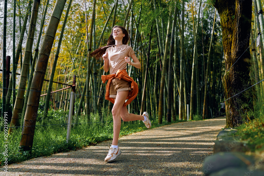 Naklejka premium Woman running along a bamboo forest path, fitness jog and outdoor exercise with a smile, casual shorts and sneakers, sunlight, motion and healthy lifestyle on a scenic trail.