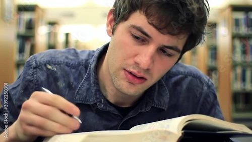 Young man studying intensely in a library, writing notes from a book.