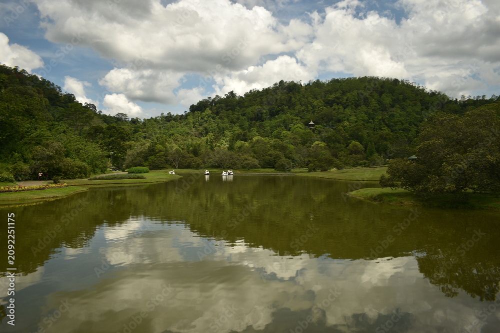 Obraz premium Scenic landscape of a calm lake reflecting fluffy white clouds and lush green rolling hills under a blue sky.