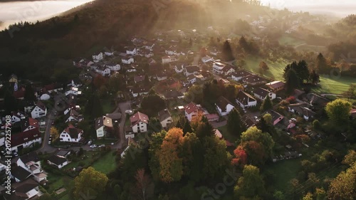Flying over a scenic village and rural landscape with hills, meadows, woods, sunbeams and illuminated fog at sunrise.