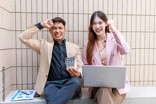 Bild auf Leinwand Stressed man with calculator and smiling woman with laptop sit by a tiled wall, mixed feelings about money, work, budgeting, or business teamwork