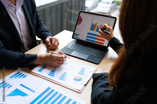 Two business professionals discussing sales charts and graphs on a laptop and paper reports during a team meeting at a desk in a modern office.