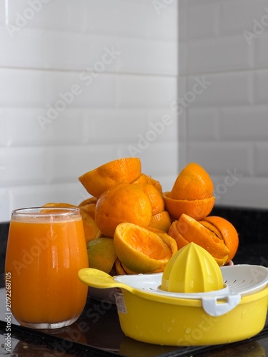 A commercial eye-level photograph of a yellow and white plastic manual citrus juicer placed in the center of many fresh orange halves, creating a neat pattern on a black reflective kitchen counter. Th