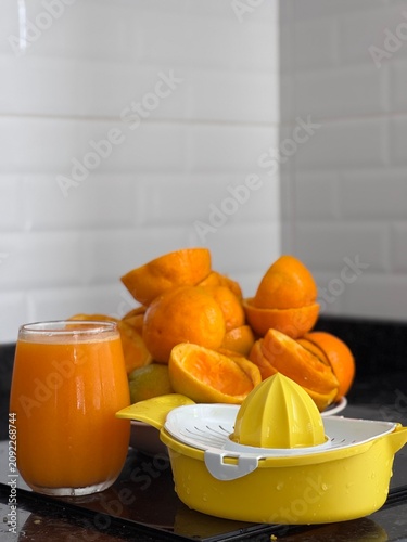 A commercial eye-level photograph of a yellow and white plastic manual citrus juicer placed in the center of many fresh orange halves, creating a neat pattern on a black reflective kitchen counter. Th