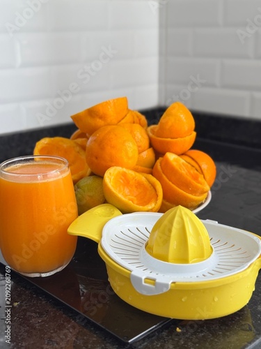 A commercial eye-level photograph of a yellow and white plastic manual citrus juicer placed in the center of many fresh orange halves, creating a neat pattern on a black reflective kitchen counter. Th