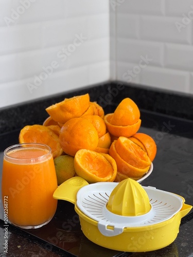 A commercial eye-level photograph of a yellow and white plastic manual citrus juicer placed in the center of many fresh orange halves, creating a neat pattern on a black reflective kitchen counter. Th