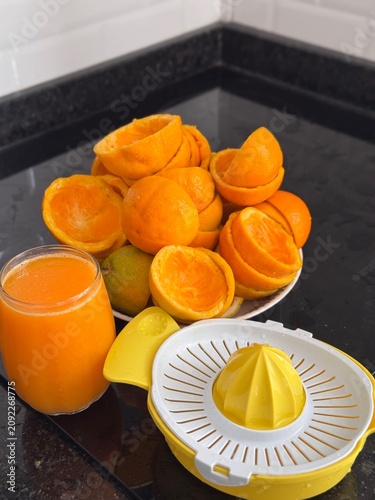 A commercial eye-level photograph of a yellow and white plastic manual citrus juicer placed in the center of many fresh orange halves, creating a neat pattern on a black reflective kitchen counter. Th