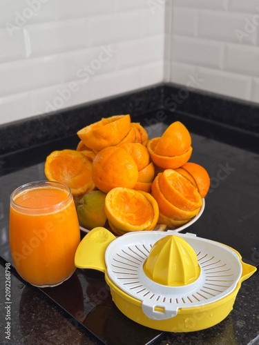 A commercial eye-level photograph of a yellow and white plastic manual citrus juicer placed in the center of many fresh orange halves, creating a neat pattern on a black reflective kitchen counter. Th