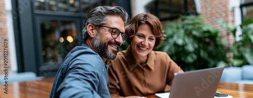 Middle-aged man and woman smiling and working together on a laptop in a modern indoor office environment with plants in the background