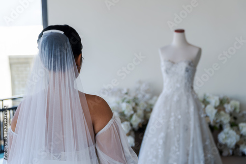 Fotografie Back view of a bride wearing a white veil looking at her beautiful wedding dress on a mannequin before the marriage ceremony