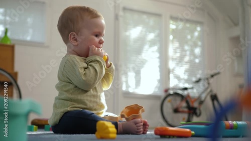 Infant sits barefoot on a colorful playmat, intently holding and chewing a yellow toy near face. Soft diffused daylight in a warm, cozy, developmentally nurturing home environment