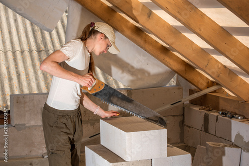 Female worker using a handsaw to cut a lightweight aerated concrete block during home renovation work on an attic construction site, focusing on efficiency and progress