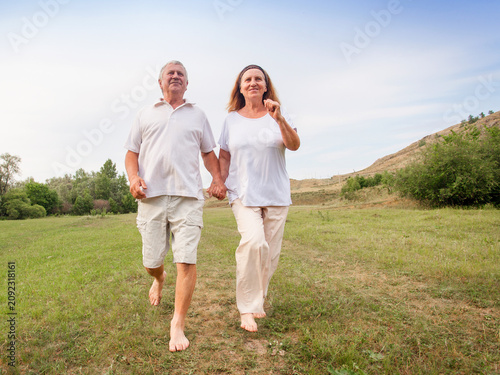 Senior man and senior woman holding hand walking barefoot on green field. Cheerful mature couple enjoying active lifestyle in nature. Concept of healthy retirement and togetherness.