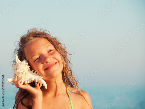 Happy child girl listening to seashell at beach. Young girl holding conch shell to her ear to hear ocean sound. Summer vacation activity, childhood holiday, leisure travel concept.