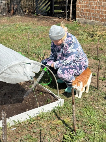 A woman waters a high raised garden bed with a hose, which is covered with spunbond over plastic arches.
