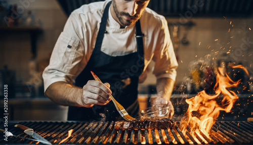 Chef grilling a steak with flames on a commercial grill.