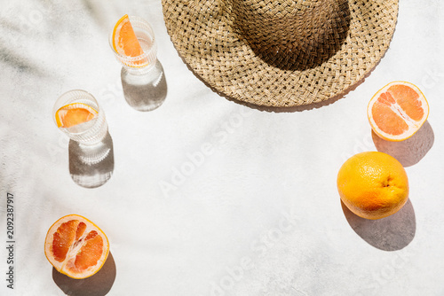 A straw hat and fresh grapefruit slices with cold glasses of water on a sunny surface under palm leaf shadows, tropical vacation vibe, refreshing lifestyle, summertime drinks.