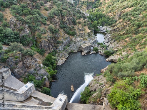 Fotografía Water flows from a dam into a river surrounded by rocky hills and dense greenery