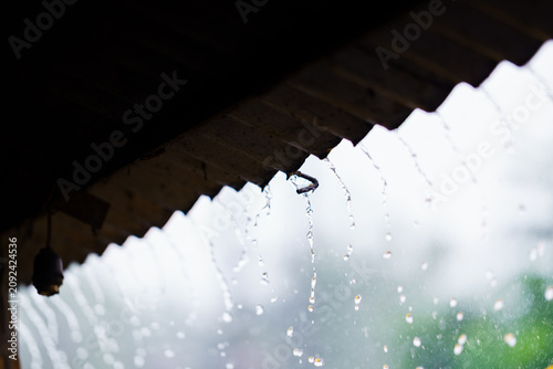 Rainwater dripping from the edge of a corrugated metal roof during a heavy downpour, creating a rhythmic pattern of falling water droplets in a rural setting