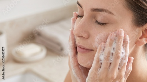 Young woman washing her face with soft white soap foam in a bright natural bathroom