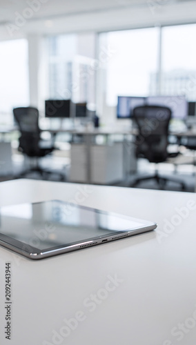 A digital tablet computer lies on a white office desk with a blurred corporate workspace background