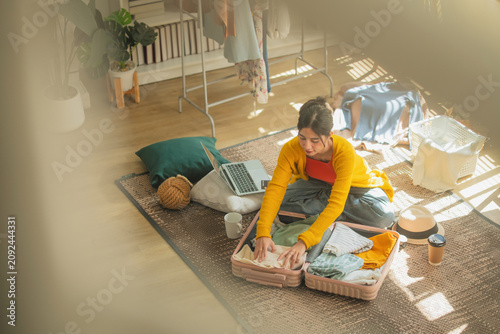 Asian woman packing clothes into open suitcase on bedroom floor surrounded by messy room, overhead view of travel preparation and trip planning lifestyle concept