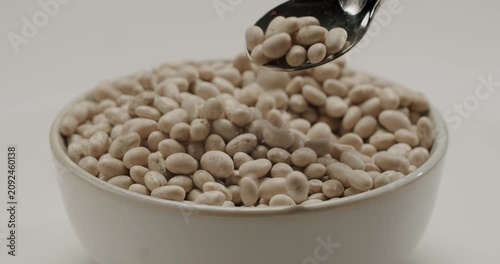 Slow motion capture of a spoon scooping raw white cannellini beans from a full ceramic bowl, with the legumes falling back, all isolated on a clean white background for a fresh concept