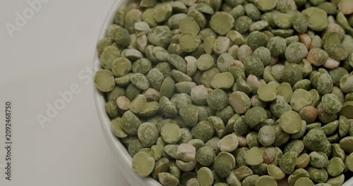 Dried green split peas in a white ceramic bowl rotating slowly. Green dried split peas rotating slowly. Spoon scooping and pouring raw dried green split peas into a white ceramic bowl
