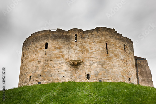 An exterior view of the historic stone Clifford's Tower (York Castle) sitting on a grassy mound under an overcast, moody sky in York, United Kingdom. HDR.