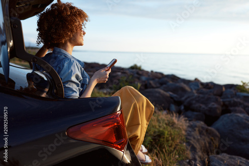 Obraz na płótnie Young woman with curly hair sits in a car trunk by a rocky shore, scrolling on a