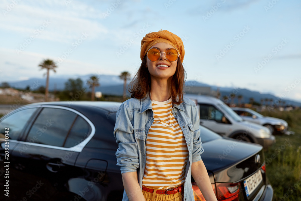 Naklejka premium Smiling woman outdoors wearing striped shirt denim jacket orange hat and sunglasses. Casual lifestyle portrait in bright daylight with a relaxed, cheerful mood near parked cars.