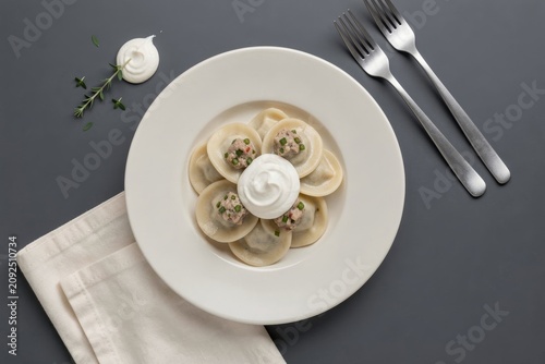Plate of pelmeni topped with sour cream, garnished with herbs, silver forks beside. Concept of dumplings with meat pepper sour cream on gray surface, appealing presentation.