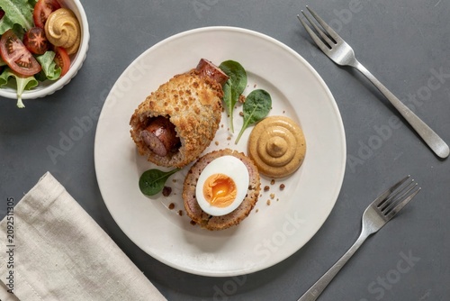 Scotch Egg cut in half with golden-brown yolk, surrounded by fresh spinach. Concept of crispy sausage wrapped egg with breadcrumbs fried on plate with fork, bowl of tomatoes.