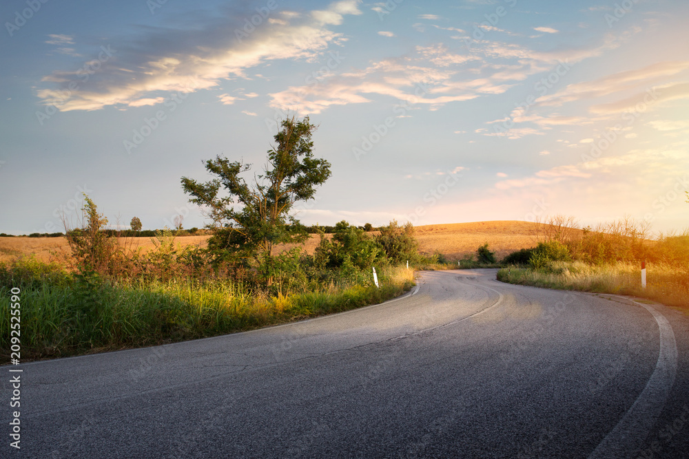 Naklejka premium Winding country road through golden field at sunset, curvy asphalt road landscape in Tuscany, Italy