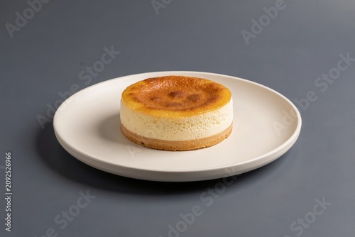 Sernik on small round white plate with creamy texture, light yellowish-brown crust, dark grey surface. Concept of baked cheesecake with twarog vanilla crust and dessert presentation.