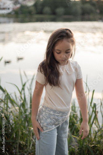 Fotografie A thoughtful young girl with long brown hair stands by a lake, wearing a light sweater and flowy blue pants, surrounded by greenery