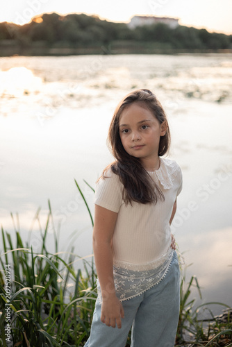 A young girl with long brown hair poses by the water's edge at sunset, wearing a light-colored top and denim pants.